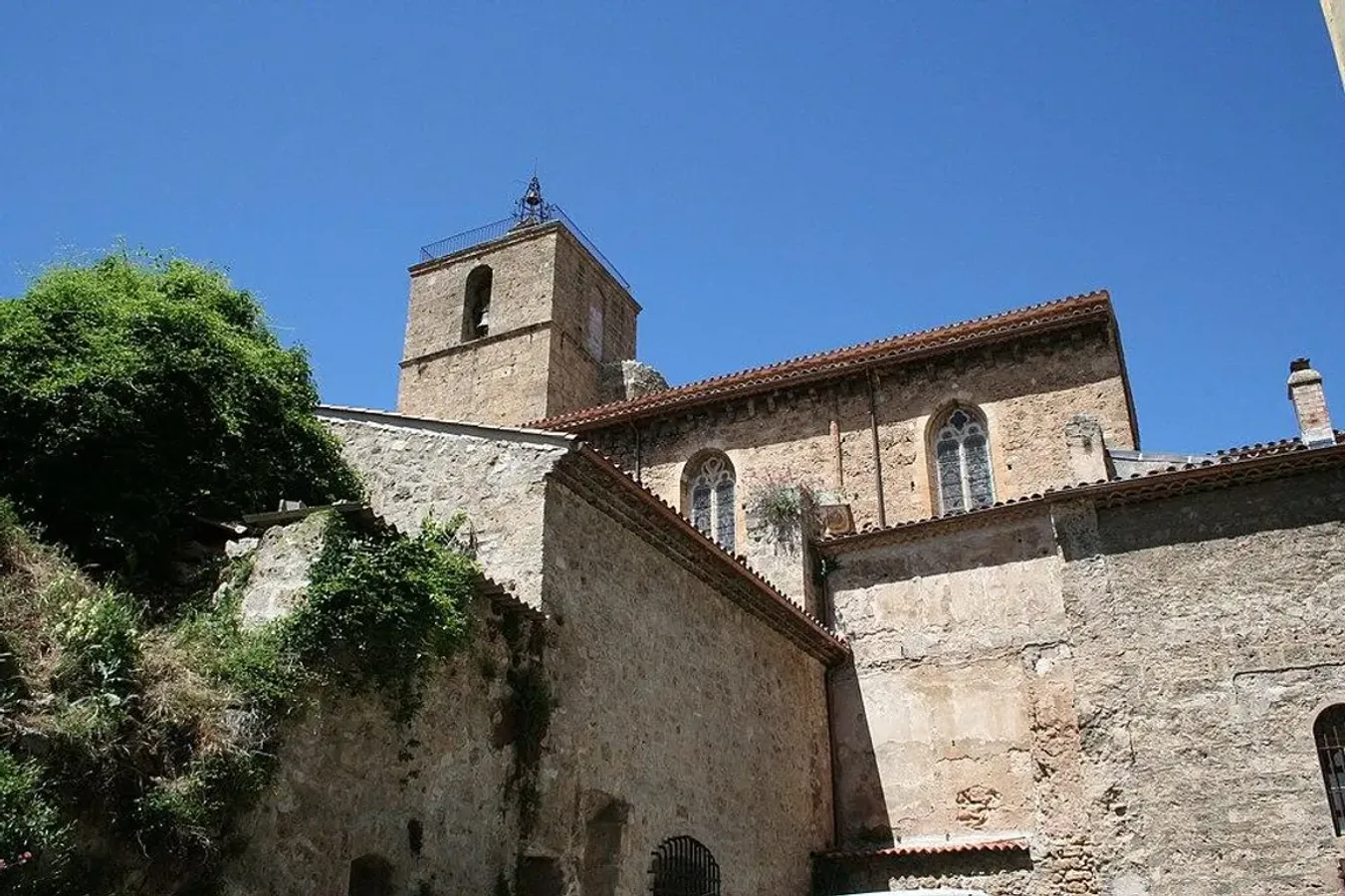 Collégiale Saint-Marcel en Provence avec un clocher en pierre s’élevant au-dessus des toits en tuiles rouges, sous un ciel bleu clair, photographiée en contre-plongée.
