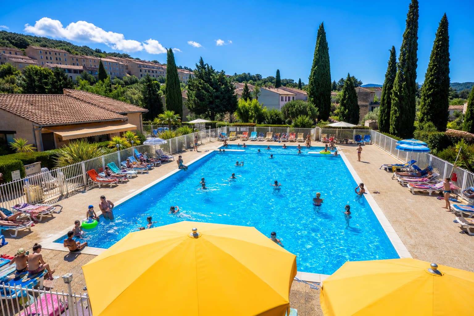 Vue aérienne d’une piscine communale animée dans un village français en été, avec une eau bleue cristalline, des transats colorés, des parasols et des cyprès méditerranéens.