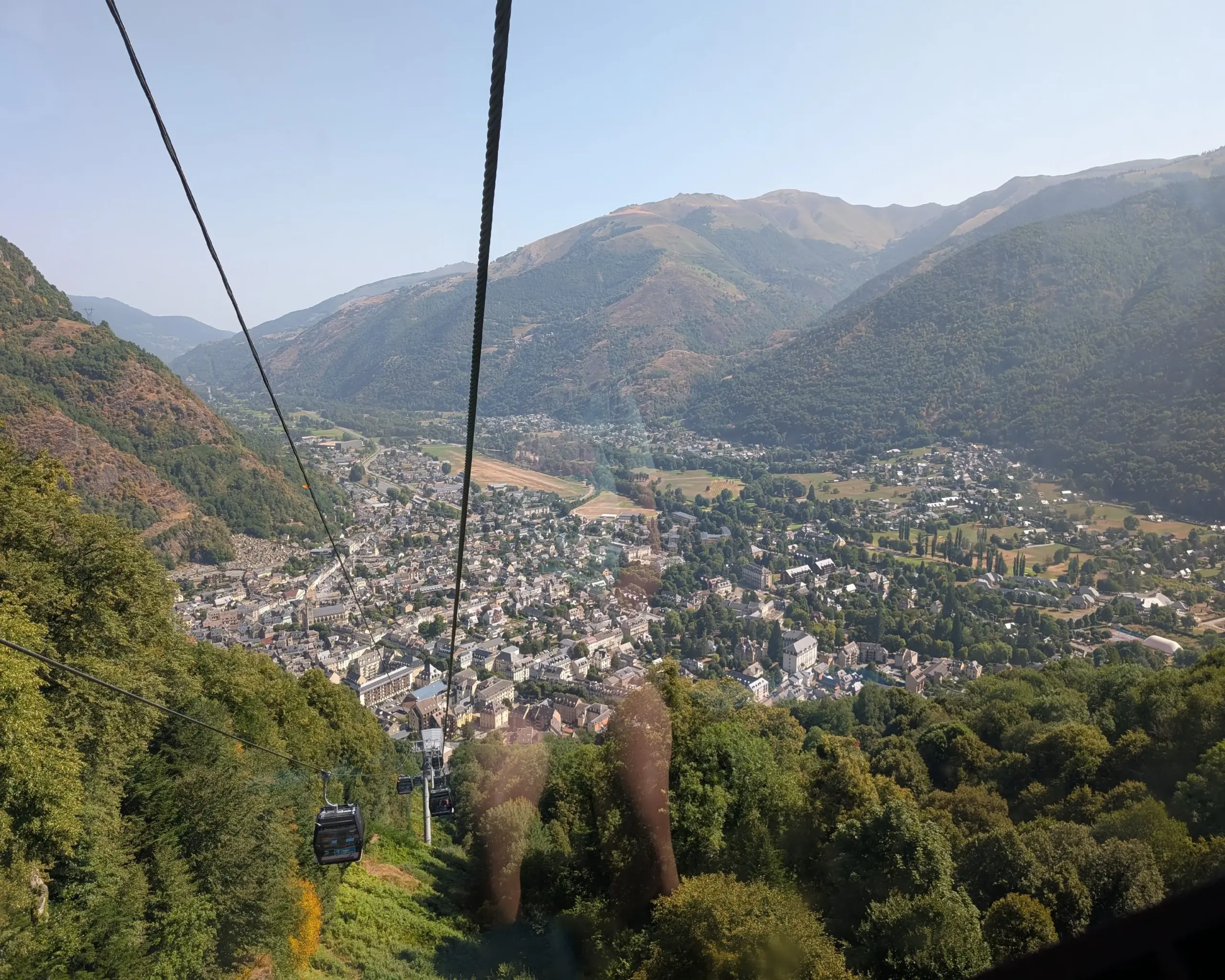 Un téléphérique survole une vallée verdoyante des Pyrénées, avec des randonneurs et vététistes sur les sentiers en contrebas, et la ville de Luchon au loin, sous des couleurs estivales éclatantes.