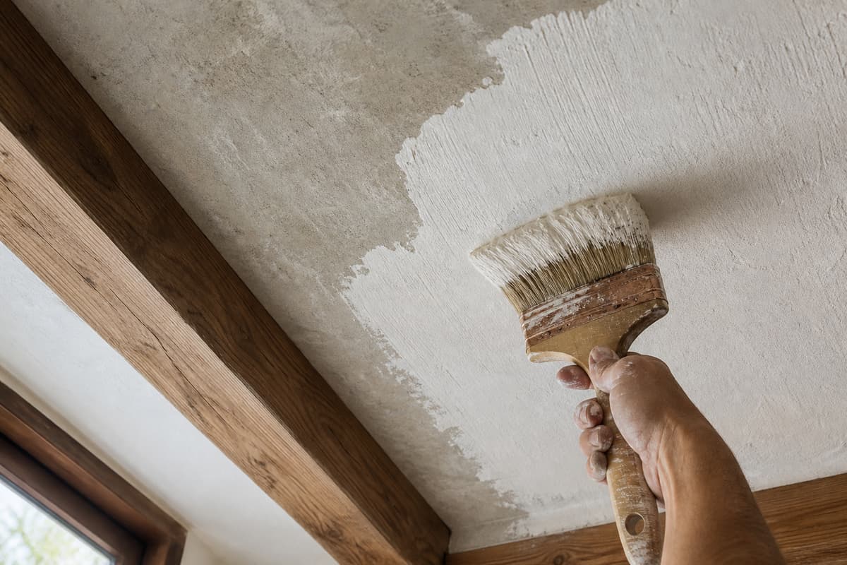 Main passant un badigeon de chaux clair sur une surface minérale de plafond entre des poutres en bois propres, avec un rendu mat légèrement irrégulier.