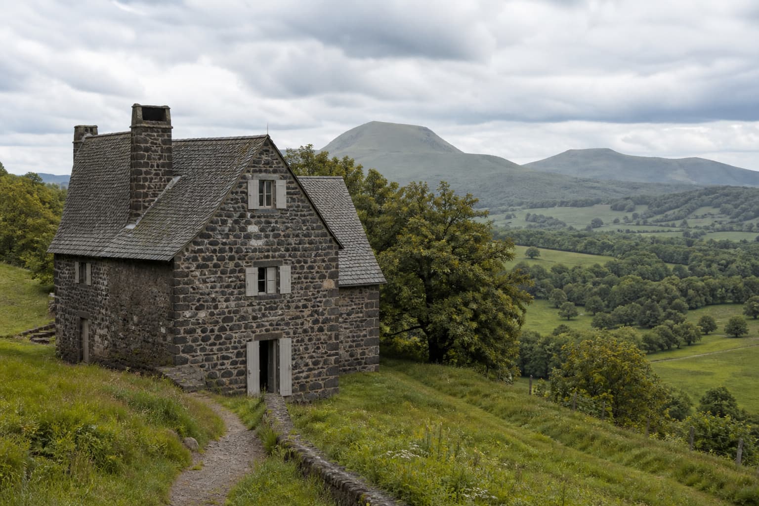 Demeure ancienne en pierre sombre avec toit traditionnel, nichée dans un paysage verdoyant et vallonné typique de l’Auvergne, avec des reliefs volcaniques à l’arrière-plan.