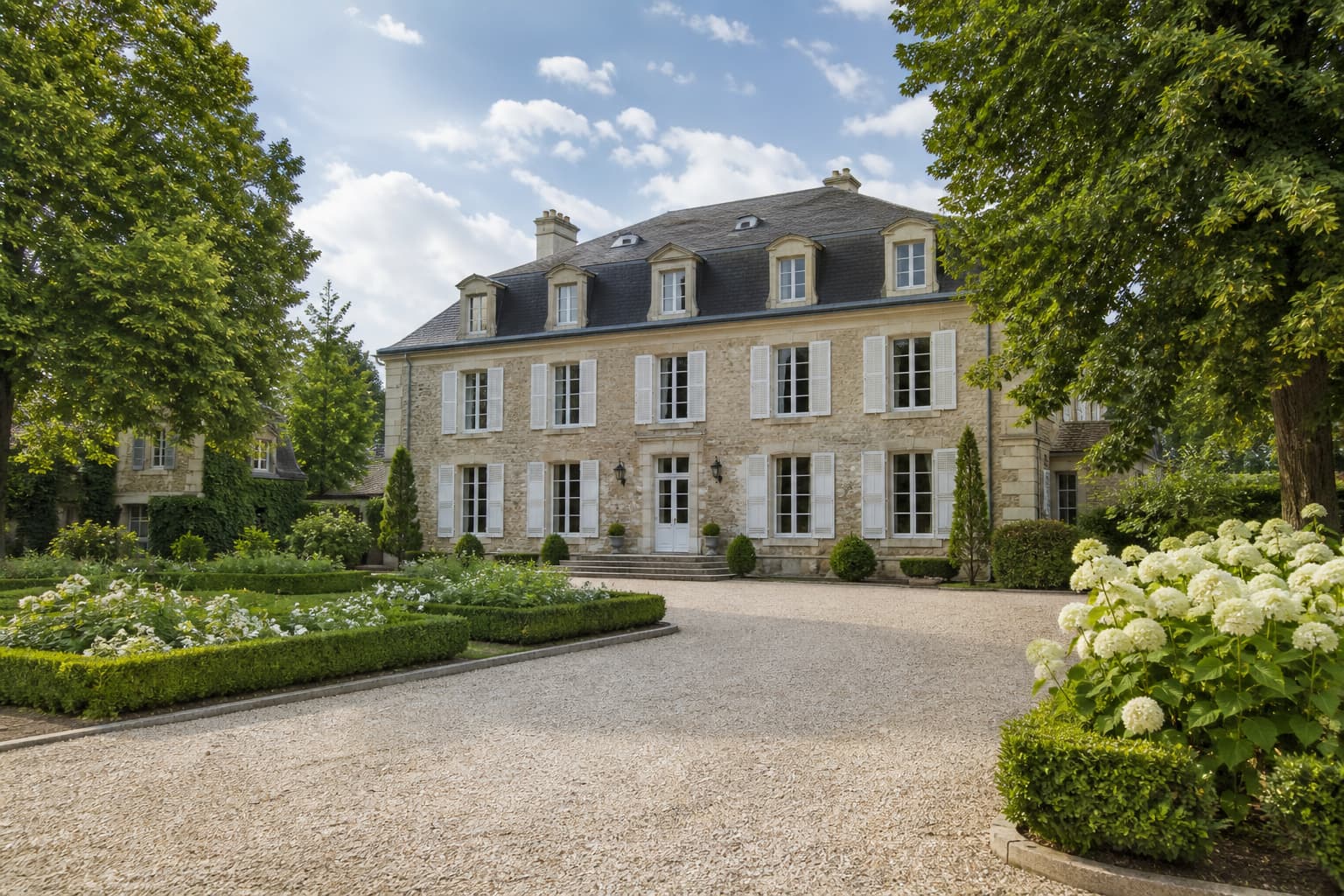 Façade d’un manoir historique en pierre avec volets blancs et jardin soigné, situé près de Paris.