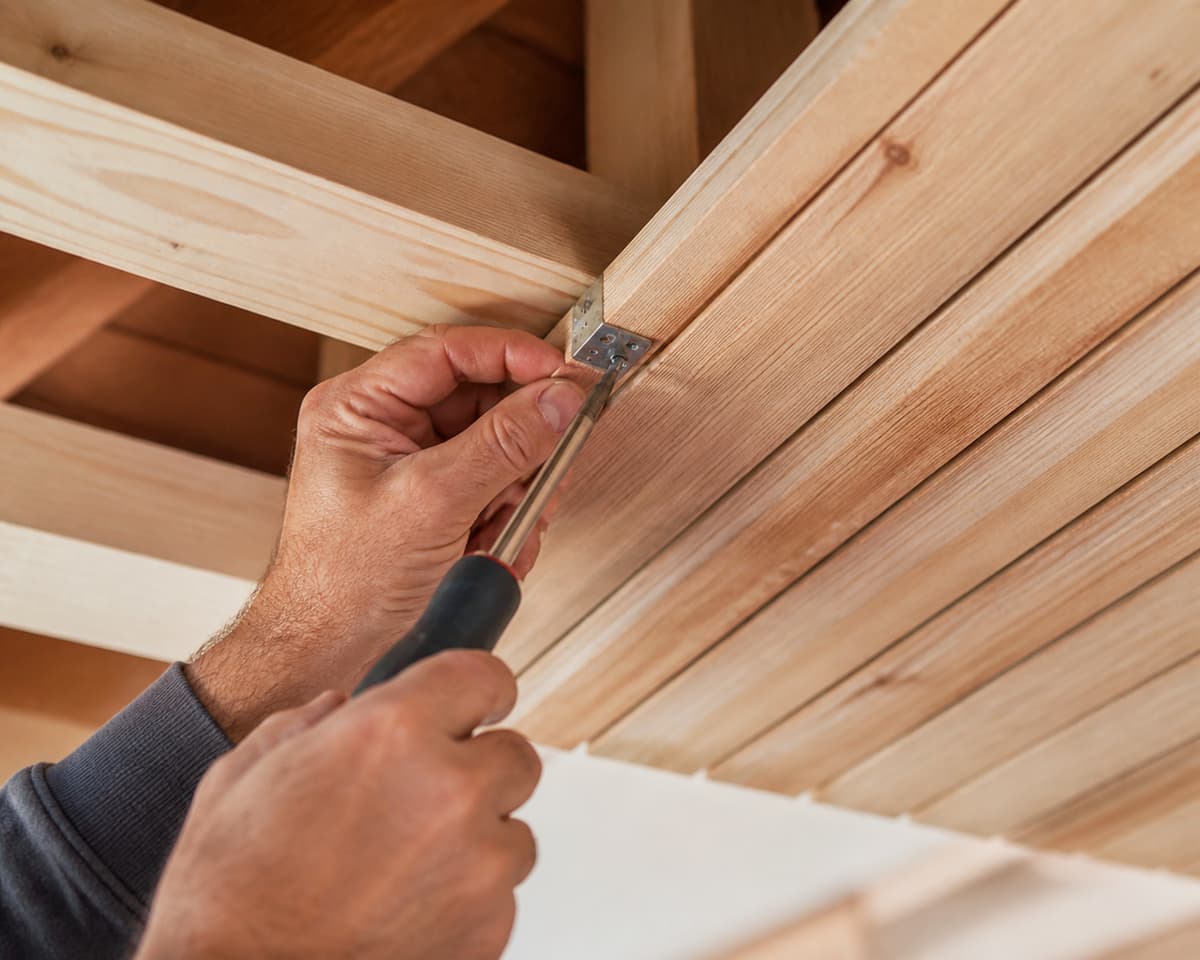 Gros plan sur des mains fixant des planches de bois alignées sur une structure de plafond, dans un chantier propre.