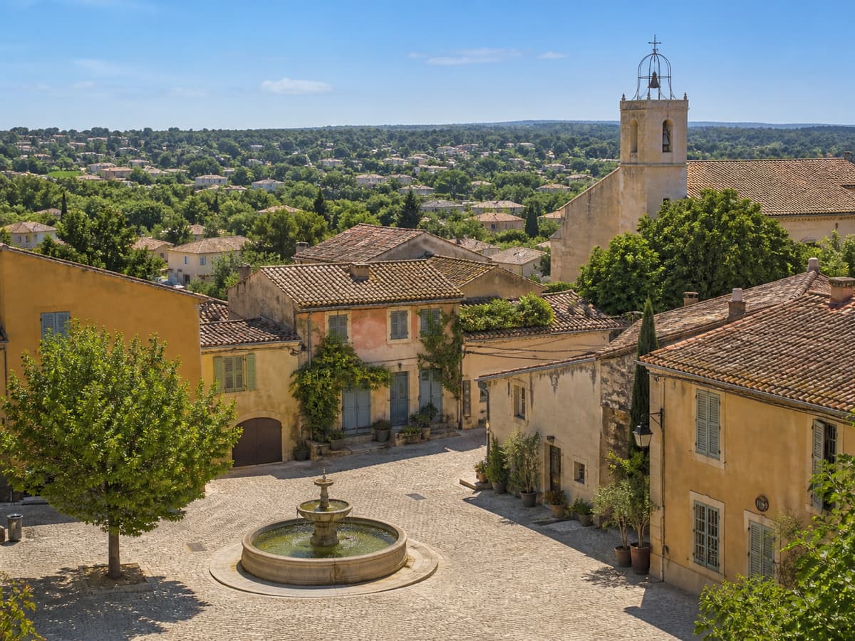 Une place de village provençal avec une fontaine en pierre, une église en arrière-plan, des maisons provençales et un paysage périurbain.